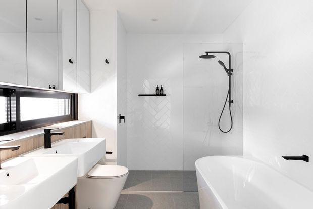 Bright contemporary bathroom with a double floating vanity, wall-mounted black tapware, integrated storage, a toilet, freestanding-style bathtub, and an open walk-in shower behind a glass screen. White cabinetry and herringbone wall tiling are balanced by a grey tiled floor and black fixtures.