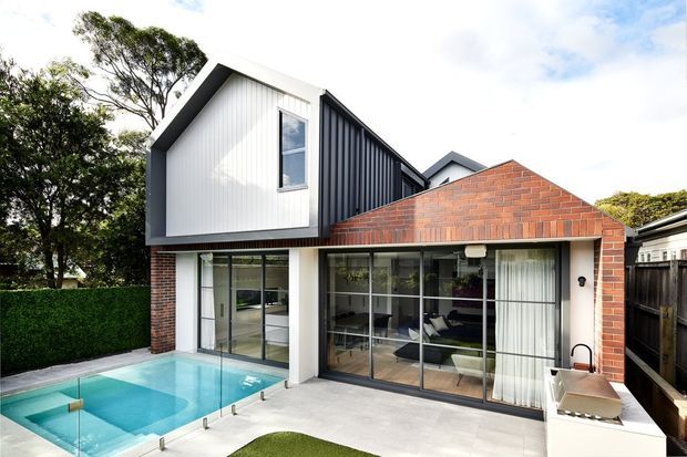 Contemporary two-storey residential extension with a bold gabled upper volume clad in dark vertical metal and light panels, set above a low brick wing with a pitched brick roof. Full-height glazed doors open to a paved courtyard and turquoise swimming pool, with a compact outdoor kitchen at the right. Mature hedging and trees frame the suburban setting.