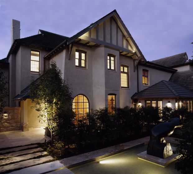 Exterior dusk view of a substantial multi-storey house with steep gabled roofs, dark tiled roofing, rendered walls, timber-framed windows, illuminated interiors, landscaped hedges, and a lit driveway or garden path. A sculptural feature sits in the foreground.