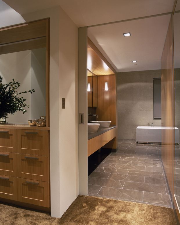 View into a refined bathroom from an adjoining vanity or dressing area. The bathroom features a freestanding white bathtub, twin vessel basins on dark stone joinery, warm timber wall panelling, large grey stone floor and wall tiles, recessed ceiling lights, and a frameless glass shower enclosure. A timber built-in cabinet and decorative planting are visible in the adjacent area.