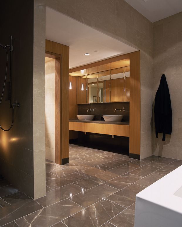 A sophisticated contemporary bathroom viewed from beside a freestanding bathtub, featuring a walk-in shower, large-format beige stone-look wall and floor tiles, a timber-lined vanity alcove with two vessel basins, dark benchtop and backsplash, mirrored cabinetry, and warm integrated lighting.