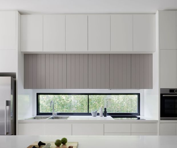 Bright, minimalist contemporary kitchen with extensive white flat-panel cabinetry, a grey vertical-grooved backsplash, a wide black-framed horizontal window, stainless-steel sink and tap, and integrated appliances. The kitchen opens toward leafy greenery outside, while a pale island or benchtop occupies the foreground.