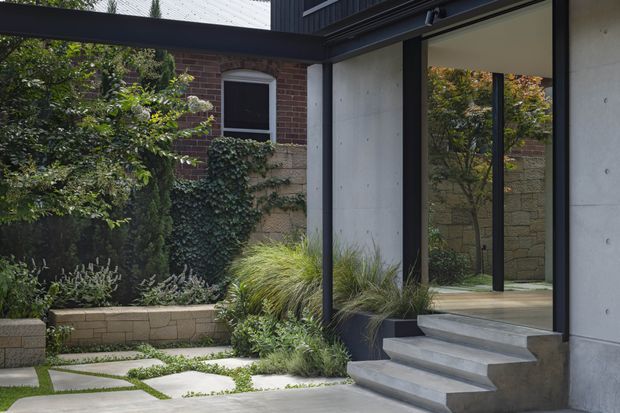 A contemporary residential entrance and landscaped courtyard, viewed from outside. The composition features a covered entry with dark slender metal framing, a large glazed door or window, smooth concrete wall panels, broad concrete steps, and a paved garden path bordered by layered planting. A red-brick building and rendered masonry boundary wall are visible in the background.
