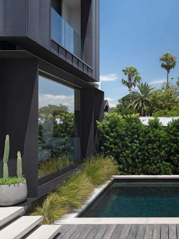 A close exterior view of a dark, angular contemporary house beside a rectangular swimming pool. The facade features charcoal cladding, large floor-to-ceiling glazing, strong cantilevered forms and deep architectural reveals. A timber deck, pale paving, ornamental grasses, hedging and potted cacti surround the pool, with palm trees visible beyond.