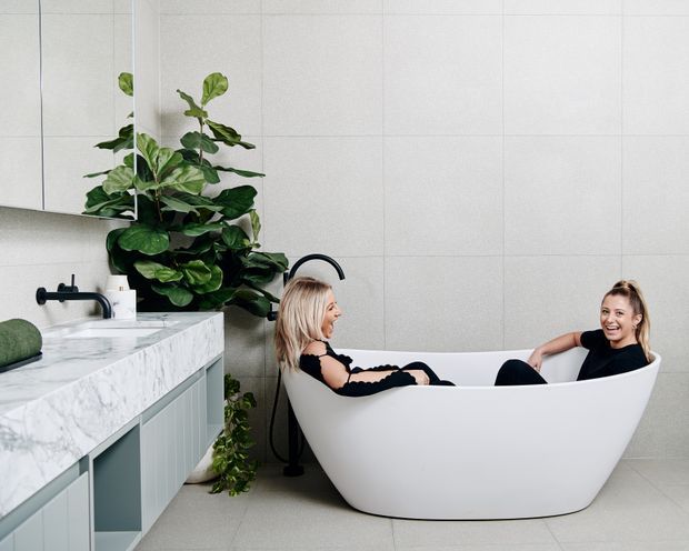 Bright contemporary bathroom featuring a freestanding white oval bathtub, pale tiled walls, a marble-look vanity countertop with blue-grey cabinetry, matte black tapware, a large mirror, and abundant indoor greenery. Two people are relaxing in the bath.