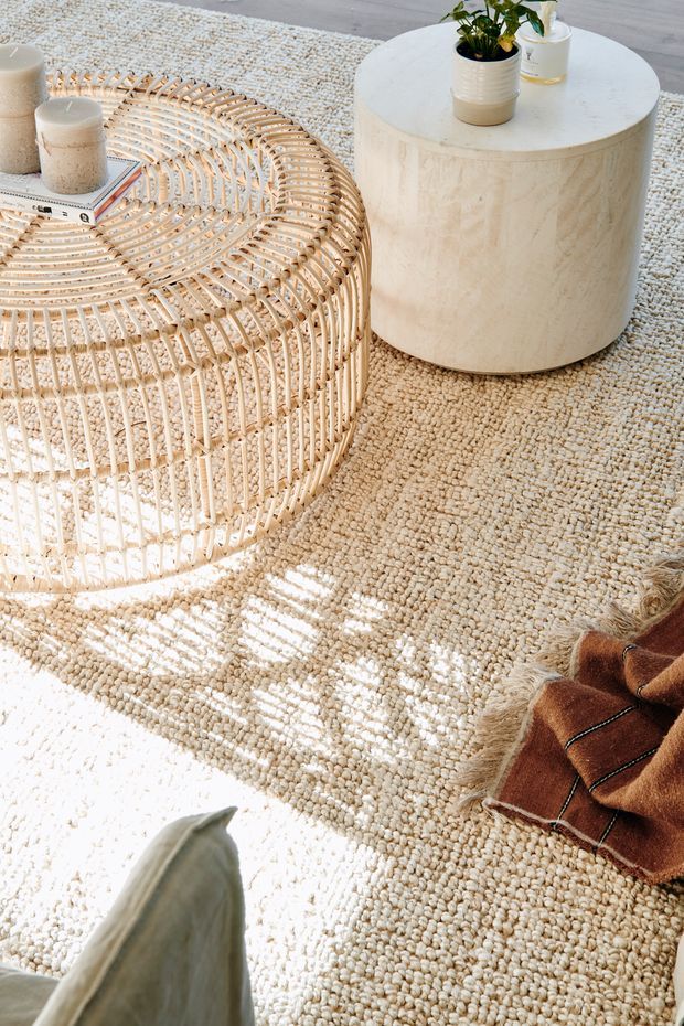 A warmly lit living room vignette featuring a large cream textured rug, a rounded woven rattan coffee table with books and candles, and a cylindrical pale stone or travertine side table topped with a potted plant. A rust-brown fringed cushion or throw sits at the lower right, creating a natural, relaxed, tactile composition.