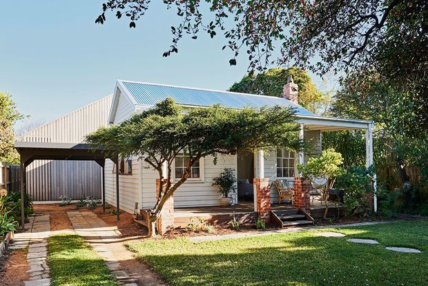 A single-storey traditional cottage-style home viewed from the front, with pale weatherboard cladding, a pitched gabled corrugated metal roof, a covered front verandah supported by simple posts, brick porch columns, and a detached or adjoining covered carport at the left. The house sits within a landscaped suburban garden with a lawn, stepping stones, mature trees, and shrubs.