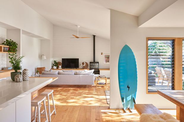 Bright open-plan contemporary interior combining a kitchen, dining area and living room. The space features light timber flooring, white walls and cabinetry, a large pale kitchen island with bar stools, built-in shelving, a low sectional sofa, and abundant natural light through timber-framed louvred glazing. A surfboard displayed near the opening adds a coastal character. The room has a warm, relaxed and airy atmosphere.
