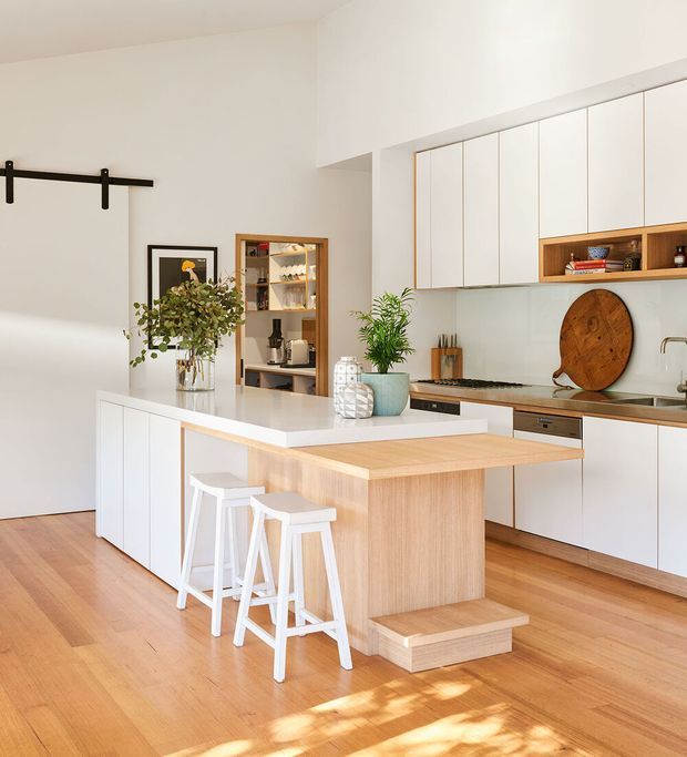 Bright contemporary kitchen featuring extensive white flat-panel cabinetry, a large white island with a light timber breakfast bar, white stools, warm timber flooring, stainless-steel appliances and hardware, and open display shelving. A black barn-style sliding door rail and framed artwork add subtle contrast. Natural light creates a warm, inviting atmosphere.