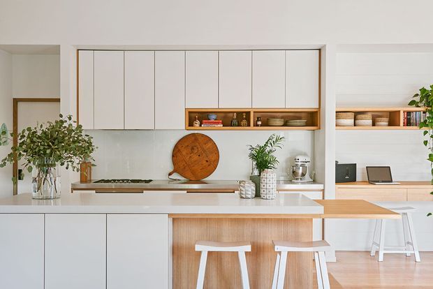 Bright, open-plan contemporary kitchen with white handleless cabinetry, a pale stone island benchtop, timber-lined breakfast bar, integrated cooktop, and open timber shelving. The space connects visually to a compact study or living area with built-in shelving and a desk. Indoor plants and warm timber accents soften the clean, light scheme.