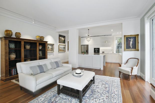 Bright open-plan living room with a white upholstered sofa, patterned area rug, dark wood coffee table, traditional display shelving, artwork, and a classic armchair. The room opens directly into a contemporary white kitchen with an island, integrated cabinetry, and pendant lighting. Polished timber flooring, white walls, detailed ceiling cornices, and large glazed doors create a refined, airy interior.