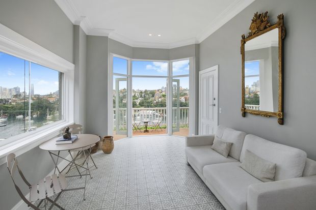 Bright, refined sitting room with a large white sectional sofa, small round dining or occasional table and chairs, patterned pale rug, grey walls, white architectural trim, and an ornate gilded wall mirror. Floor-to-ceiling bay-style glazing and a balcony open to a leafy urban waterfront outlook.