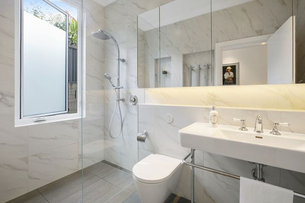 Bright contemporary bathroom featuring full-height white marble-look wall tiling, grey stone-look floor tiles, a frameless glass shower enclosure with chrome fixtures, wall-mounted toilet, and a large white double-ended basin with exposed chrome plumbing. A wide mirrored cabinet and concealed warm LED lighting run above the vanity. The room has a clean, refined, spa-like appearance with a frosted window providing natural light.