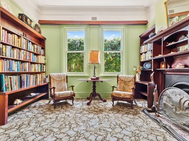A traditional, richly detailed library or reading room with floor-to-ceiling dark timber bookshelves, pale green walls, ornate white ceiling mouldings, two sash windows, a small round pedestal table, upholstered antique armchairs, patterned carpet, and a decorative fireplace with a mirrored overmantel. The room feels warm, refined, and heritage-inspired.