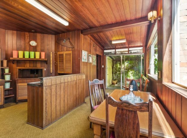 A warm, wood-lined dining and living area with extensive timber wall and ceiling panelling. The room includes a timber dining table and chairs in the foreground, a built-in timber bar or counter with louvred cabinet doors, open shelving with colourful pottery, and a fireplace or dark recessed niche at the rear. Large glazed doors and windows open toward a leafy outdoor area, bringing in natural light. The space has a distinctly retro, richly timbered character.