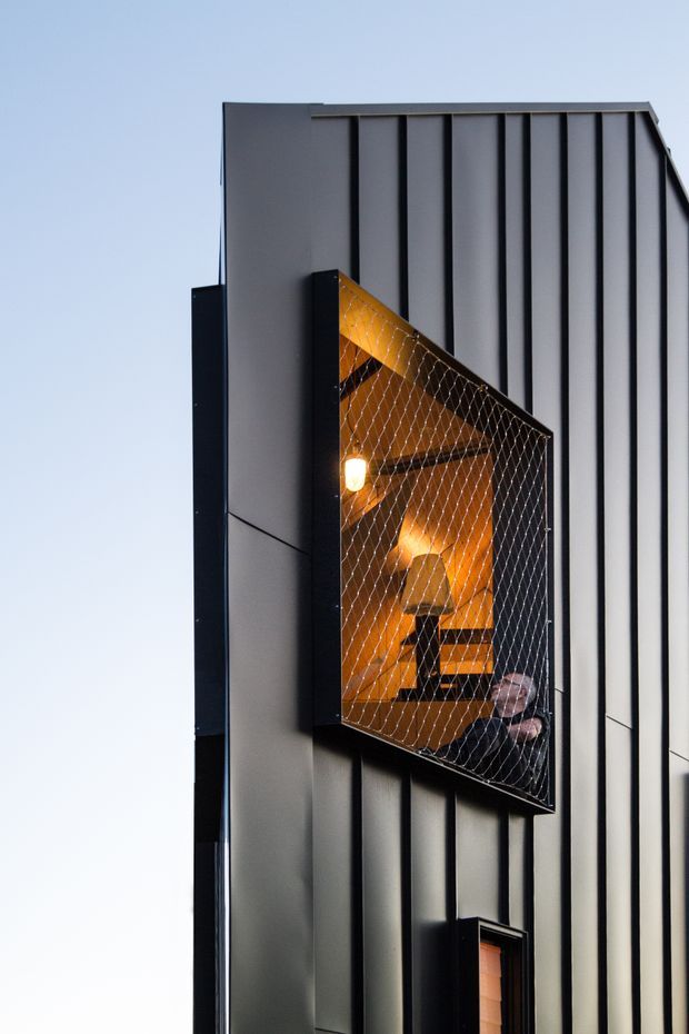 Vertical photograph of a contemporary multi-storey building facade clad in dark grey vertical metal panels. A projecting box-like balcony or window enclosure with wire-mesh screening reveals a warmly lit interior with seating and a pendant light. The composition emphasizes strong vertical ribbing, black framing, geometric projection, and contrast between the cool exterior and amber interior light.