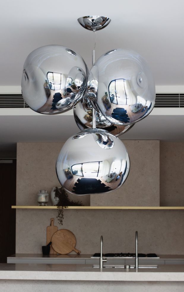 Close-up view of a contemporary kitchen centred on a sculptural pendant light with multiple polished chrome spherical shades. Behind it are large grey stone-look wall panels, a dark cooktop, timber serving boards, stainless-steel tapware and a pale stone island benchtop. Reflections in the highly polished light shades show the surrounding kitchen and windows.