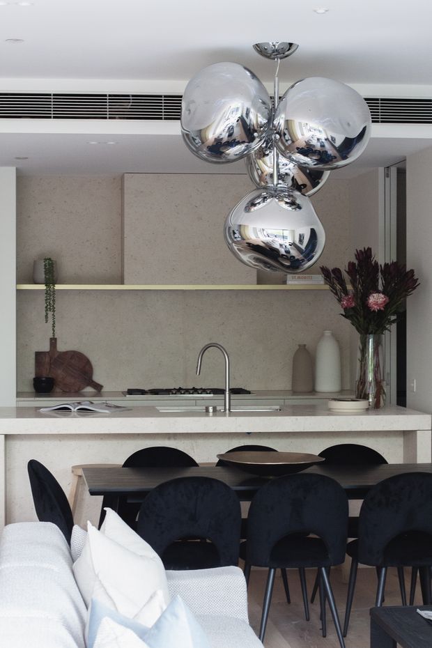 Contemporary open-plan kitchen and dining area with a pale stone island and splashback, integrated cooktop and sink, dark wood dining table, upholstered dark dining chairs, and a light sofa in the foreground. Reflective chrome globe pendant lights hang above the island, while a narrow shelf, neutral ceramics, greenery, and dried florals add restrained decorative detail.