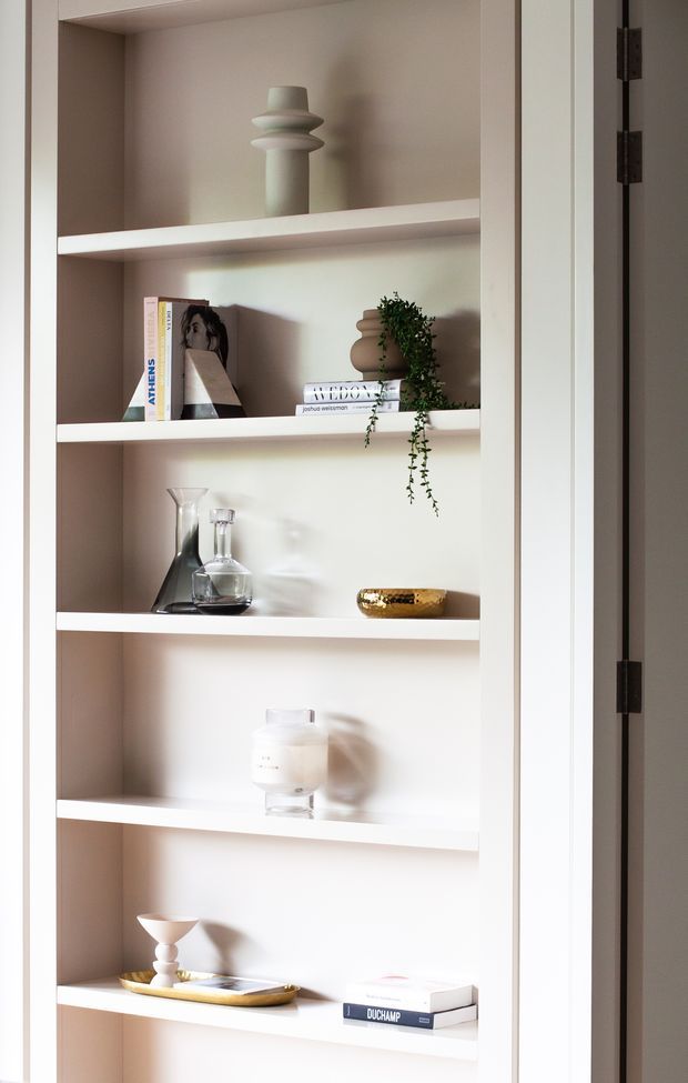 A close-up view of a built-in white painted shelving unit with multiple open shelves, styled with neutral-toned decorative ceramics, books, glass vessels, a trailing plant, and a small gold-toned tray. The shelving is set within a wall recess and framed by a white hinged cabinet or doorway panel. Soft natural light creates gentle shadows, giving the composition a calm, refined, minimalist feel.