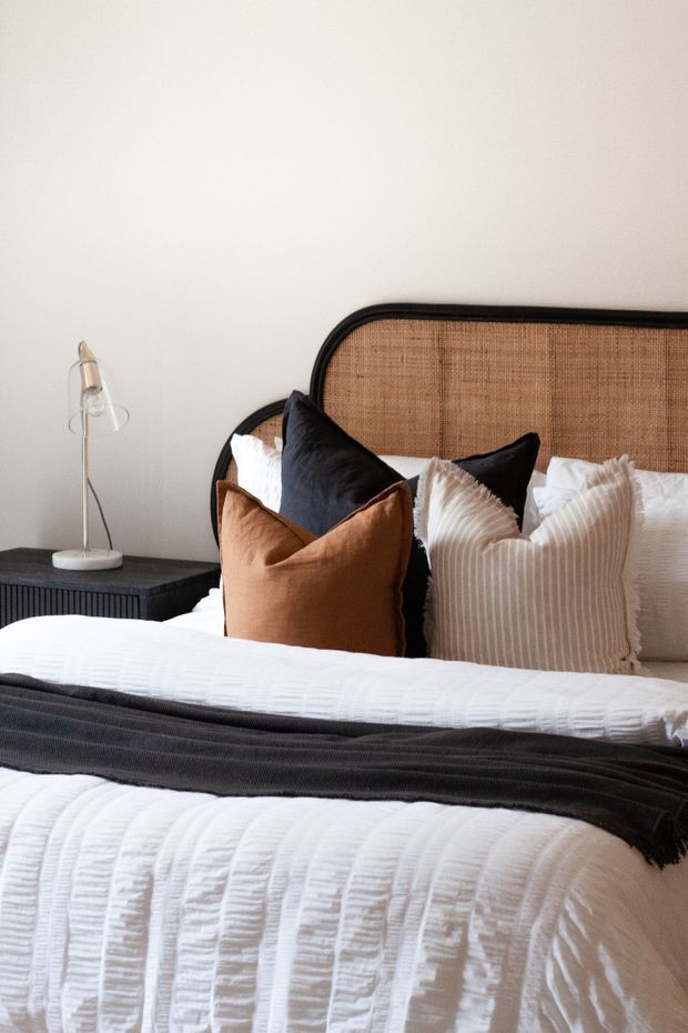 A warmly styled bedroom featuring a large bed with textured white bedding, layered black, rust-brown and cream pillows, and a dark charcoal throw. The bed has a distinctive rounded black frame and woven natural cane/rattan headboard. A small dark bedside table with vertical ribbed detailing and a slim modern lamp sits to the side. The overall look is contemporary with rich textural contrast and a neutral, earthy palette.