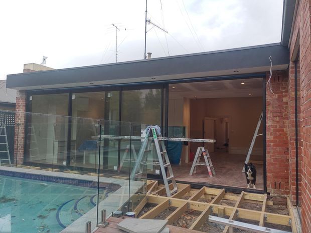 A partially completed residential renovation viewed from an outdoor pool area, showing a modern flat-roof extension with extensive dark-framed floor-to-ceiling glazing opening onto a newly constructed timber deck. Exposed brick walls, construction materials, ladders and tools are visible, with an interior room beyond the glazing.