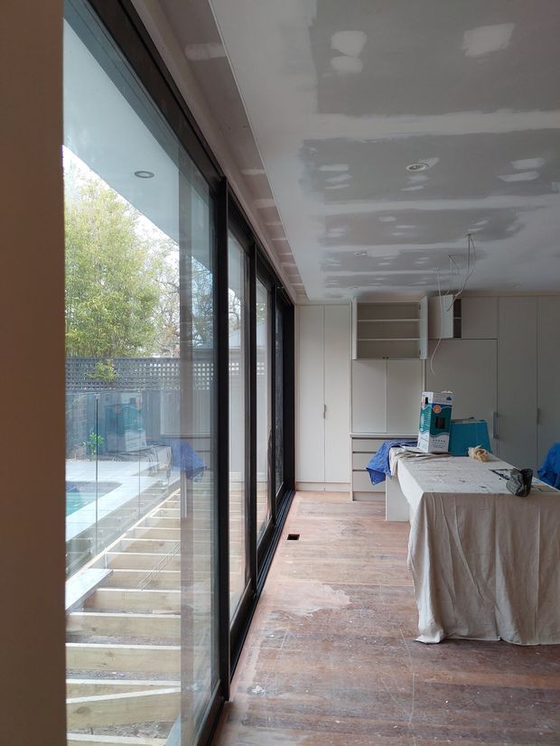 A partially finished contemporary interior under construction, likely an open-plan kitchen or living area. The room has extensive floor-to-ceiling black-framed glazing along one side, pale built-in cabinetry at the far end, unfinished plasterboard ceilings with recessed light fittings and visible joint compound, and a timber-look floor being prepared. A covered island or worktable occupies the foreground. Through the glazing, an exterior pool or courtyard area is visible.