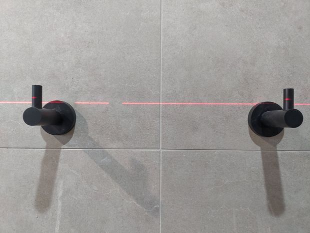 Close-up of two matte black wall-mounted bathroom fixtures or controls against large-format light grey stone-look tiles. A thin horizontal red laser line crosses the tiled wall, and the fixtures cast shadows downward.
