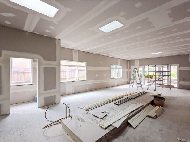 A large, unfinished interior under construction, with plasterboard walls and ceiling awaiting sanding and painting. Multiple skylights and windows provide natural light, while construction materials, ladders, scaffolding, and tools are spread across the concrete floor. The open-plan space appears to be a future living or multipurpose room.