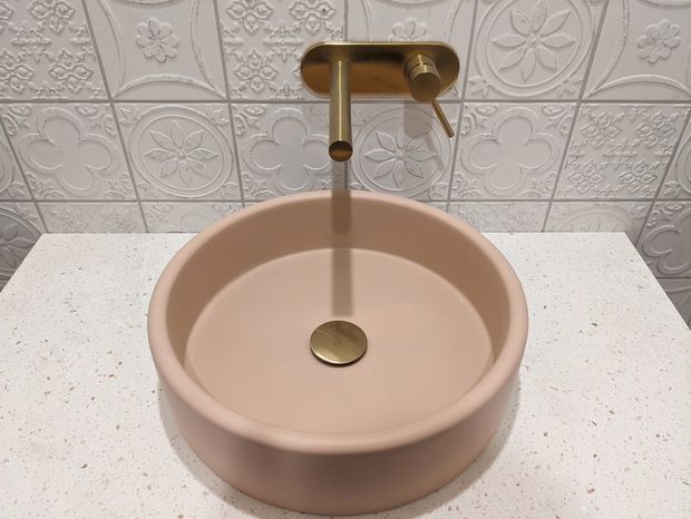 Close-up of a round blush-pink vessel bathroom basin on a light speckled benchtop, paired with a brushed brass wall-mounted tap and drain. The wall behind is clad in white embossed decorative tiles with floral and geometric motifs.