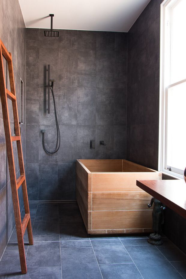 A compact contemporary bathroom featuring a freestanding timber-clad bathtub, large dark grey wall and floor tiles, a black ceiling-mounted rainfall shower, and a handheld shower rail. A tall window provides strong natural light, while a timber ladder-style shelf and dark timber vanity edge add warmth.