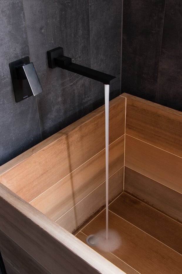 Close-up of a contemporary bathroom featuring a deep built-in bathtub lined with warm wood-look panels, dark charcoal textured wall tiles, and matte black wall-mounted bath fixtures. A slim white water stream flows from the angular black spout into the tub.