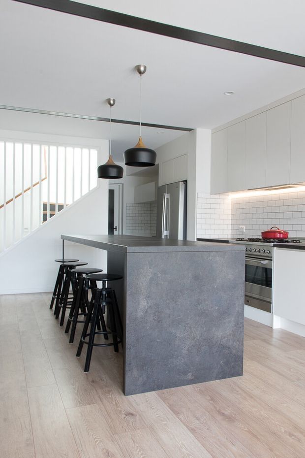 Bright contemporary kitchen with a large dark grey island and integrated seating for several people. White flat-panel cabinetry, a white subway-tile splashback, stainless-steel appliances, pale timber flooring, black pendant lights, and a staircase with white vertical balustrades are visible.