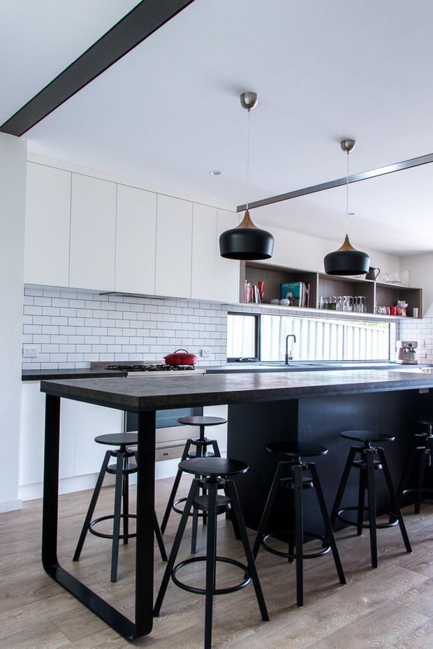 A bright contemporary kitchen centred on a large black island with an integrated breakfast bar and multiple black stools. White flat-panel cabinetry lines the rear wall above a white subway-tile splashback, while dark stone-look benchtops, black pendant lights, open shelving, stainless fixtures and light timber flooring create a crisp monochrome palette with warm flooring accents.