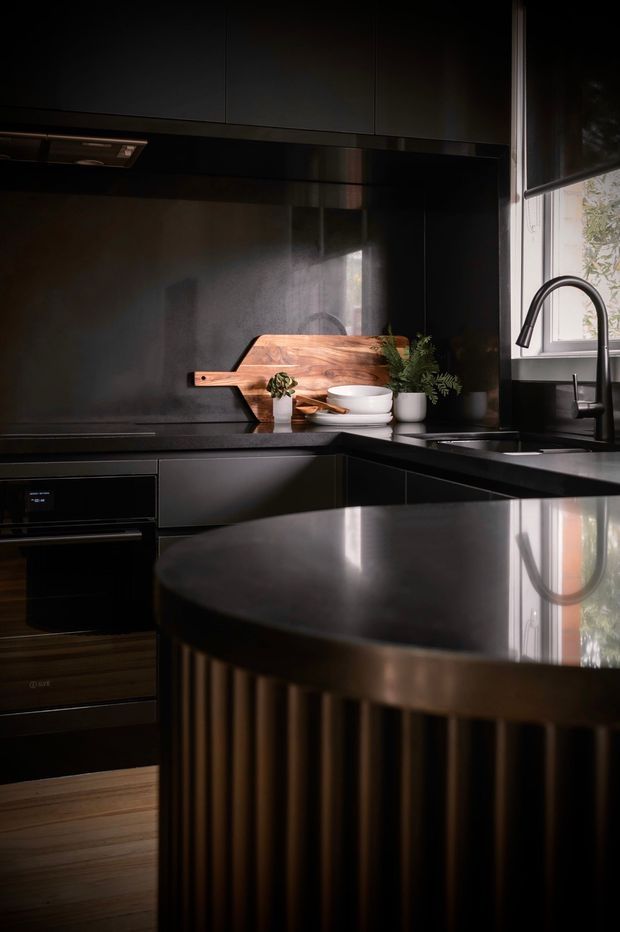 A moody, compact contemporary kitchen in a dark palette, featuring matte black cabinetry, a dark reflective benchtop and splashback, integrated black appliances, a deep black sink with a curved gooseneck tap, and a warm timber cutting board. A curved island with vertical fluted detailing dominates the foreground, while a window brings in soft natural light.