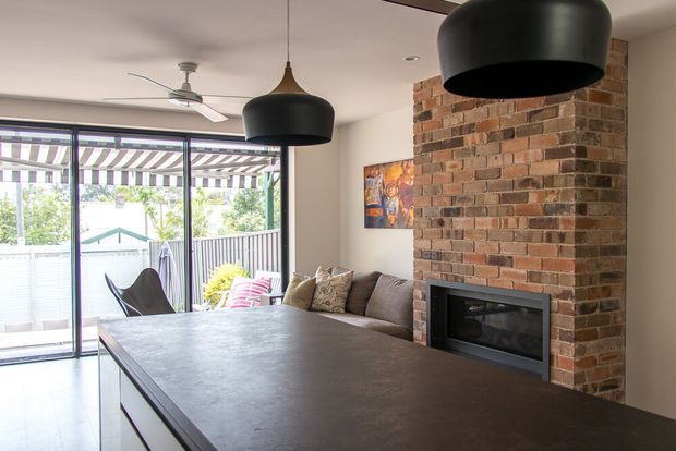 Open-plan contemporary living and kitchen area with a large dark island bench in the foreground, a neutral sectional sofa, exposed red-brown brick fireplace feature wall, black pendant lights, ceiling fan, and expansive sliding glass doors opening to an outdoor area.