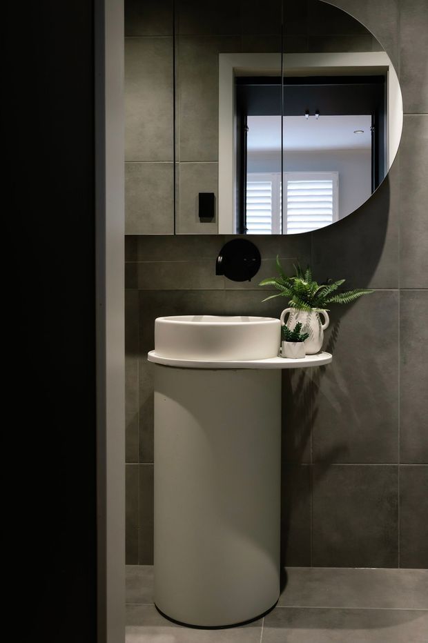 A compact, moody contemporary bathroom featuring a cylindrical white basin pedestal with a round vessel sink, dark grey tiled walls and floor, black tapware, and a large circular mirror. A small fern and ceramic vessel add a natural accent. The mirror reflects a bright window with shutters in the adjacent area.