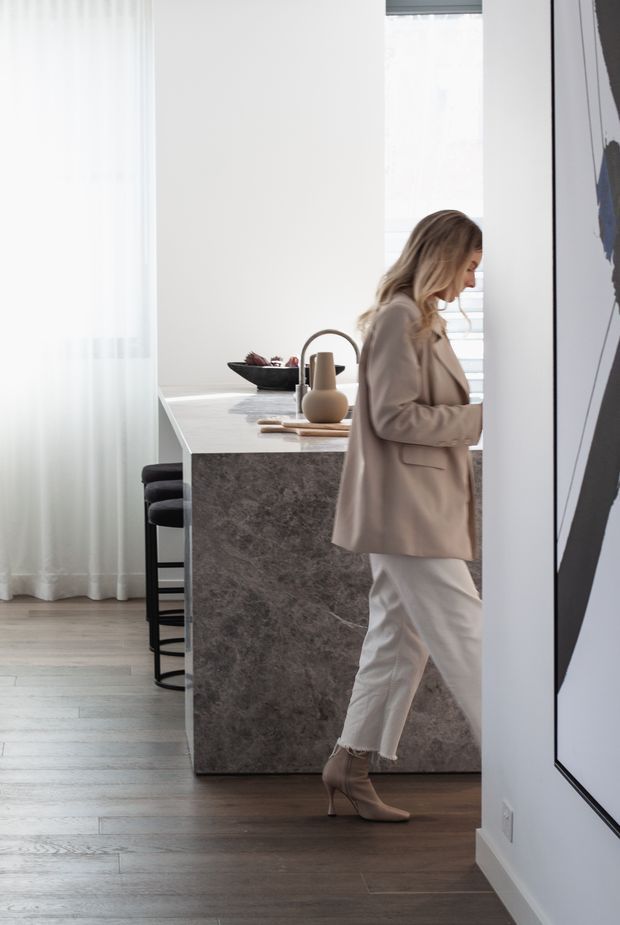 Bright, refined contemporary kitchen interior featuring a substantial stone-clad island with dark bar stools, a curved black tap, neutral accessories, sheer floor-to-ceiling curtains, pale timber flooring, and large abstract wall art. A person is walking through the foreground.