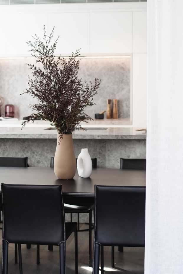 A refined contemporary dining area adjoining a kitchen. A dark dining table is surrounded by black upholstered dining chairs, with neutral ceramic vases and leafy branches as a centrepiece. Behind it, the kitchen features a grey stone-look splashback and benchtop, white cabinetry, open shelving, and soft integrated lighting. The palette is predominantly white, grey, black, and beige with a clean, understated feel.