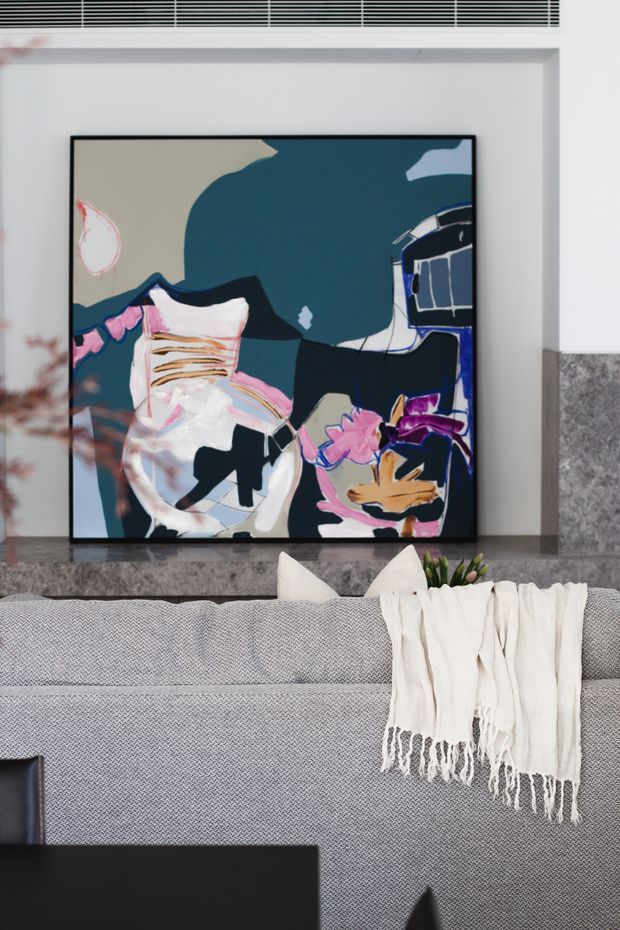 Close-up interior detail of a contemporary living room, centered on a large colourful abstract artwork above a grey textured sofa. A cream fringed throw and cushions add softness, while a dark stone or tile feature wall and white architectural detailing frame the composition.