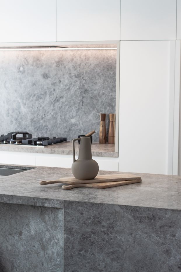 A refined contemporary kitchen featuring a large grey stone island with waterfall sides, matching stone benchtops and splashback, white flat-panel cabinetry, and a built-in gas cooktop. A sculptural dark ceramic jug and wooden cutting boards add understated styling.