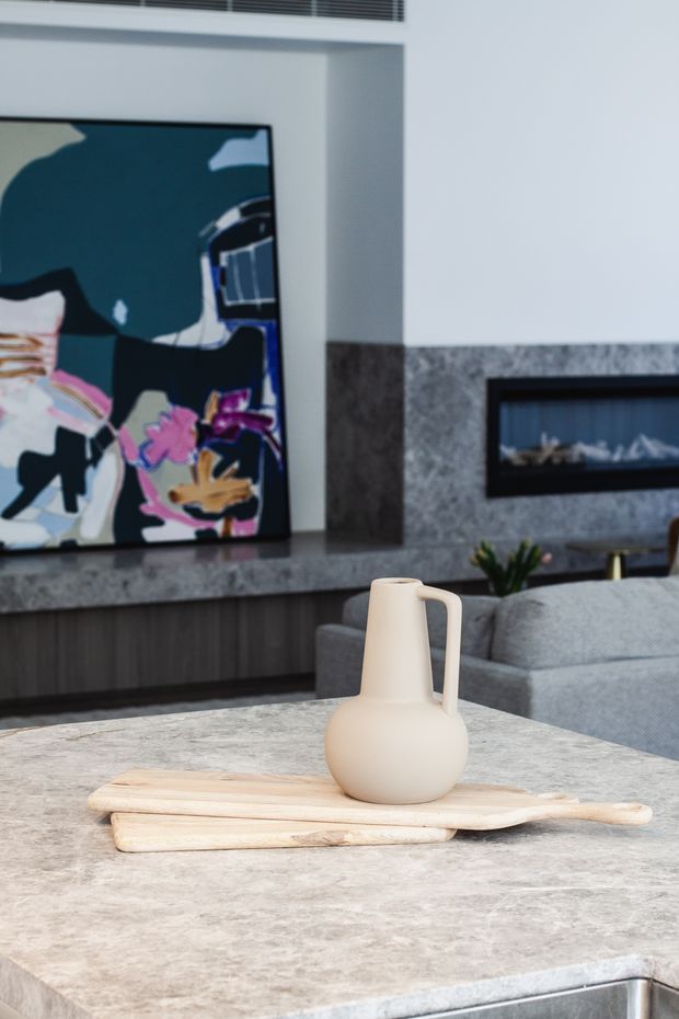 A contemporary living room viewed from behind a large stone kitchen island or benchtop. The room features grey upholstered seating, dark timber built-in cabinetry, a large abstract artwork, a linear recessed fireplace, and a grey stone-clad wall. A pale ceramic vase sits on layered wooden boards in the foreground.