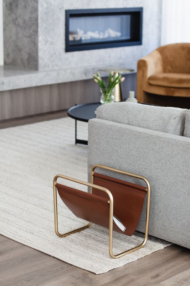 A refined contemporary living room featuring a grey upholstered sofa, black round coffee table, tan leather lounge chair, and a brown leather sling chair with brass-toned tubular frame. The room has warm grey timber-look flooring, a large light neutral rug, dark built-in cabinetry, and a textured grey stone fireplace surround with a black inset fireplace.