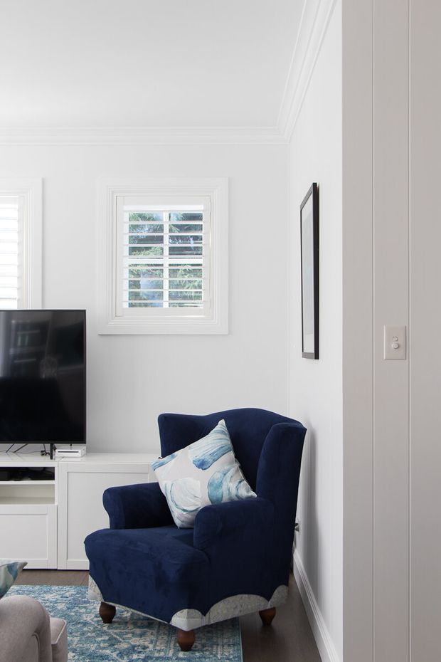 Bright, compact living room corner with white walls, detailed crown moulding and trim, plantation-shuttered windows, a navy upholstered armchair with blue-and-white cushion, patterned area rug, wall-mounted artwork, and a television on white built-in cabinetry. The space has a refined traditional feel with a cool blue-and-white palette.