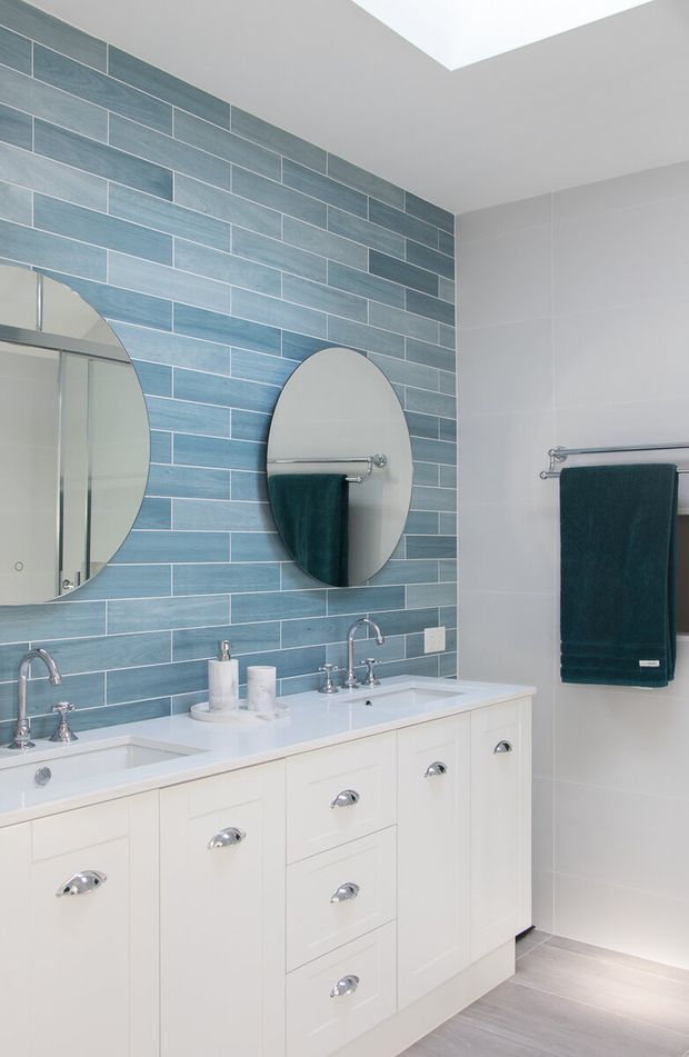 Bright, contemporary bathroom featuring a long double vanity with white shaker-style cabinetry and chrome cup pulls, twin basins, oval mirrors, chrome tapware, and dark teal towels. A blue-grey horizontally laid tiled feature wall contrasts with pale wall tiles and light flooring. A skylight provides strong natural illumination.