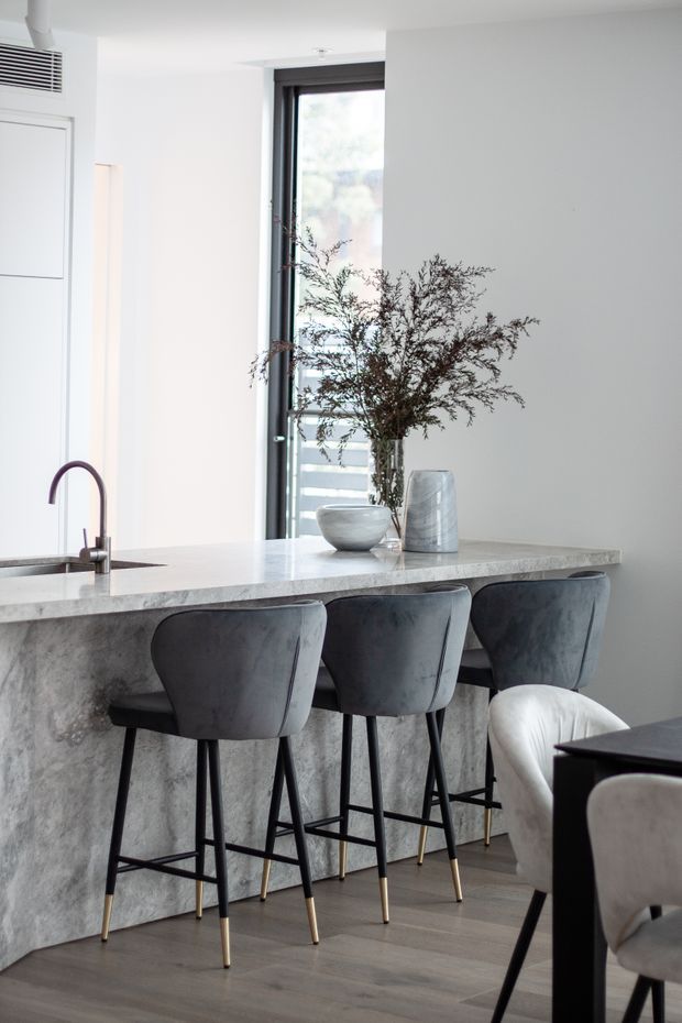 Bright contemporary kitchen interior with a long pale stone island and integrated sink, accompanied by three dark upholstered bar stools. A tall black-framed window, minimalist white walls, grey timber-look flooring, and restrained decorative styling create a refined neutral palette. Part of a dining area is visible at the right edge.