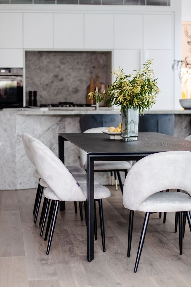 A refined open-plan dining and kitchen interior. A black rectangular dining table is surrounded by sculptural upholstered dining chairs, with a tall leafy floral arrangement as a centrepiece. Behind it is a contemporary kitchen with pale panelled cabinetry, a large grey stone island and backsplash, integrated appliances, and a dark cooktop area. Pale timber flooring and a soft neutral palette create a polished, modern atmosphere.
