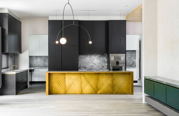 Contemporary open-plan kitchen dominated by a large central island with a striking gold-toned geometric front. The space features dark vertically grooved cabinetry, grey stone-look splashbacks and benchtops, integrated appliances, pale timber flooring, and sculptural black pendant lighting. A green built-in cabinet is visible at right.