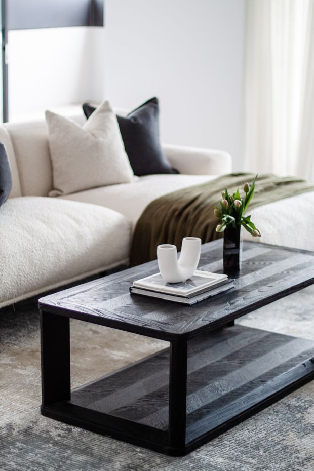 A softly lit contemporary living room featuring a large cream sectional sofa with neutral and charcoal cushions, a dark wood rectangular two-tier coffee table, and a muted patterned rug. Minimal white ceramic vessels and a small leafy arrangement sit on stacked books, while sheer curtains provide bright natural light.