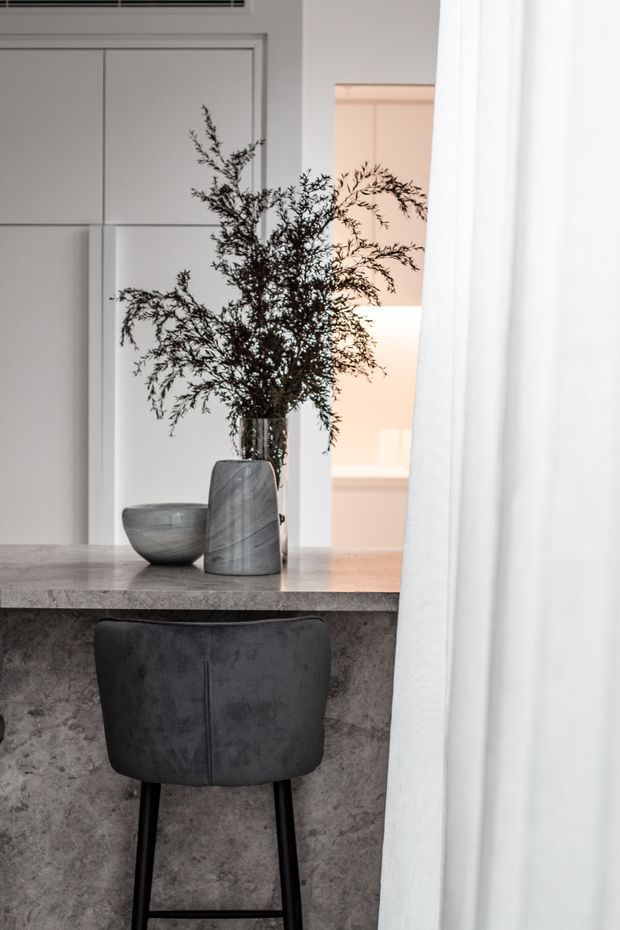 A refined interior detail showing a dark upholstered bar stool at a stone-topped peninsula or counter. The counter front has a mottled grey stone or concrete finish, with grey stone vessels and leafy branches arranged on top. White built-in cabinetry and a softly lit adjoining space are visible behind, partially screened by a sheer white curtain.