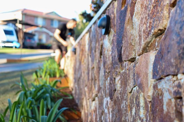 Close-up exterior view of a rough stacked-stone retaining or boundary wall, with a small black wall light mounted on the surface. A planted garden bed and residential street are visible in the blurred background.